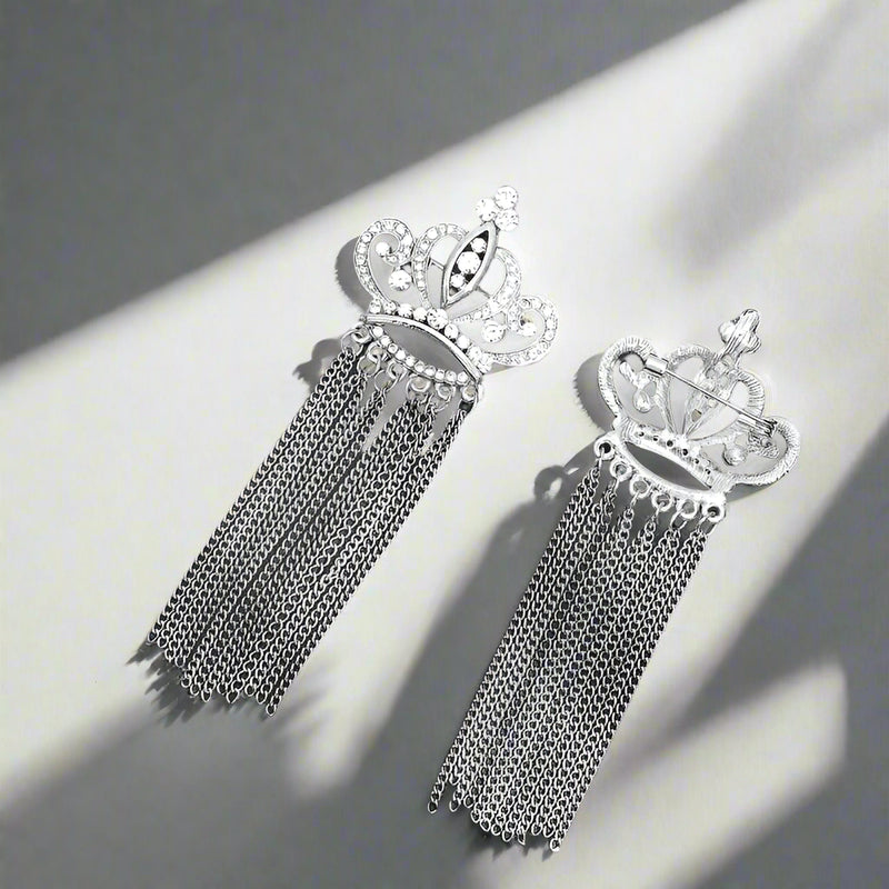 The Otre Rive Silver Tassel Brooch by Desiree W.A.R Store is showcased on a dark green backdrop with leafy shadows, featuring a crown shape, intricate designs, and rhinestone-accented dangling chains.