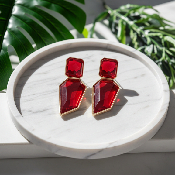 A pair of geometric drop earrings with faceted red gemstones in gold-tone settings, displayed on a round white marble tray with green leaves in the background.