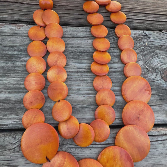 Double-strand orange wooden bead necklace with circular discs, displayed on rustic wooden background.
