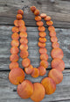 Double-strand orange wooden bead necklace with circular discs, displayed on rustic wooden background.
